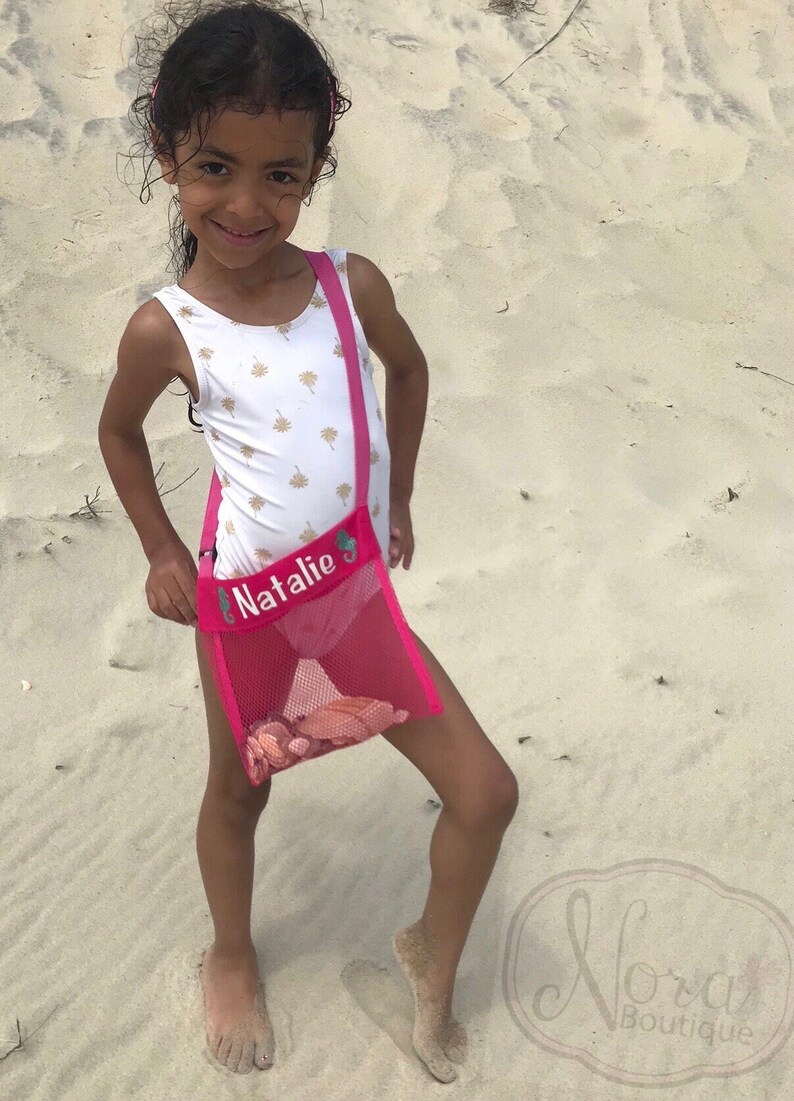 May include: A young girl wearing a white swimsuit with gold palm tree print is holding a pink mesh beach bag with the name "Natalie" embroidered on it. The bag is filled with seashells.