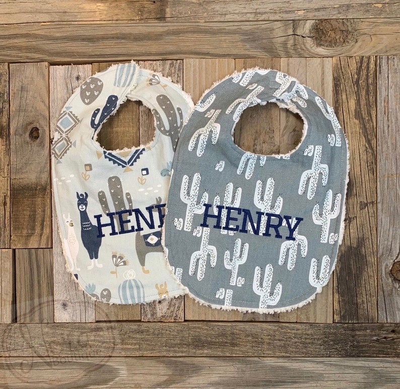 May include: Two baby bibs with a gray and white cactus print and a gray and white llama print. Both bibs have the name "HENRY" embroidered on them.