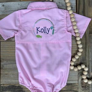 May include: A light pink baby romper with short sleeves and a collared neckline. The back features an embroidered fishing-themed design with the name "Kolly" in script. The romper is made of a soft fabric.