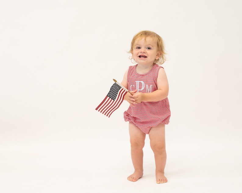 Op de afbeelding: Een jong kind draagt een rood-wit gestreept rompertje met de letters "CPM" geborduurd op de voorkant, houdt een kleine Amerikaanse vlag vast.