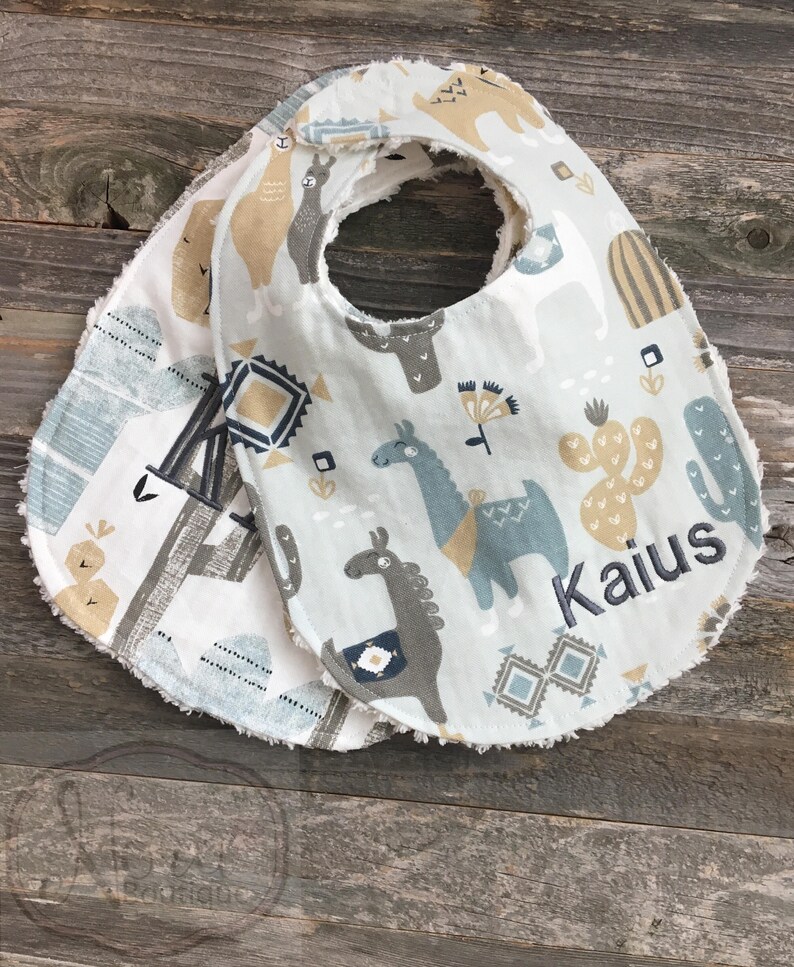 May include: Two white baby bibs with a blue and brown llama pattern. The bibs have a frayed edge and are personalized with the name "Kaius".
