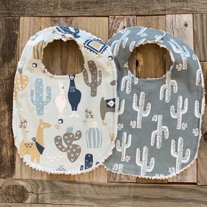 May include: Two baby bibs with a cactus and llama pattern. One bib is light blue with brown cacti and llamas. The other bib is gray with white cacti.