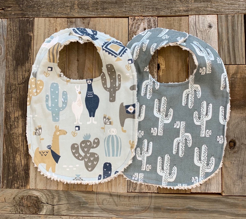 May include: Two baby bibs with a cactus and llama pattern. One bib is light blue with brown cacti and llamas. The other bib is gray with white cacti.
