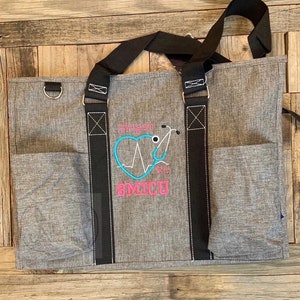 May include: Gray tote bag with black straps and a large embroidered design. The design features a pink heart with a stethoscope and an EKG line, the text "Megan RN", and the text "6MICU".