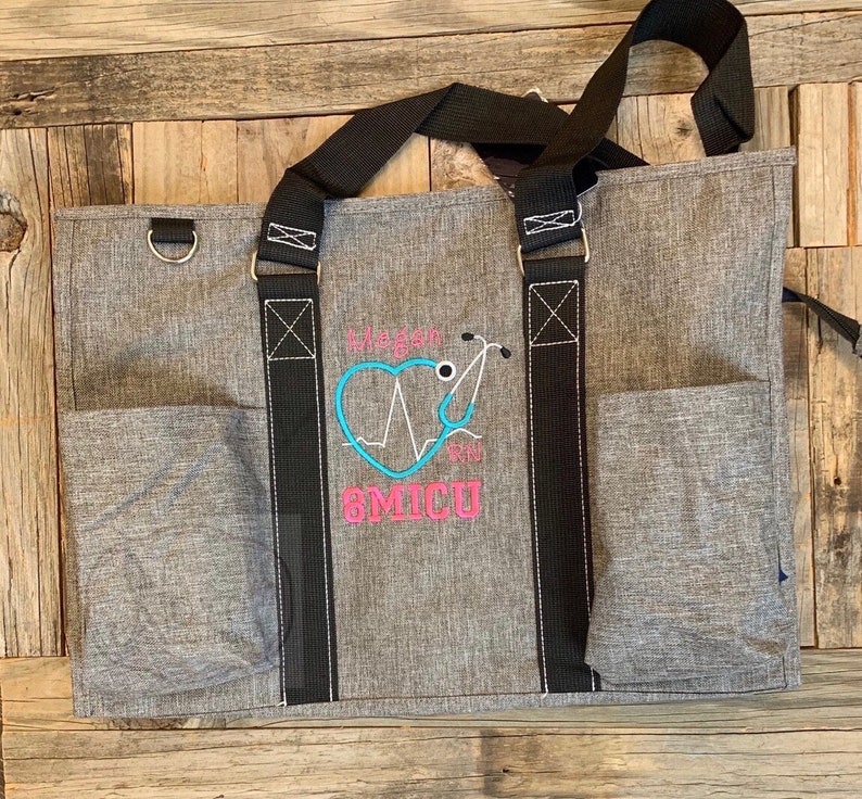 May include: Gray tote bag with black straps and a large embroidered design. The design features a pink heart with a stethoscope and an EKG line, the text "Megan RN", and the text "6MICU".