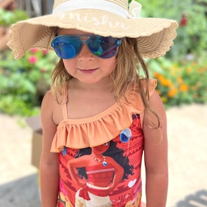 May include: A young girl wearing a straw sun hat with the name "nisha" embroidered on the brim, and blue sunglasses. She is also wearing a peach-colored swimsuit with a ruffle detail and a graphic of a character.