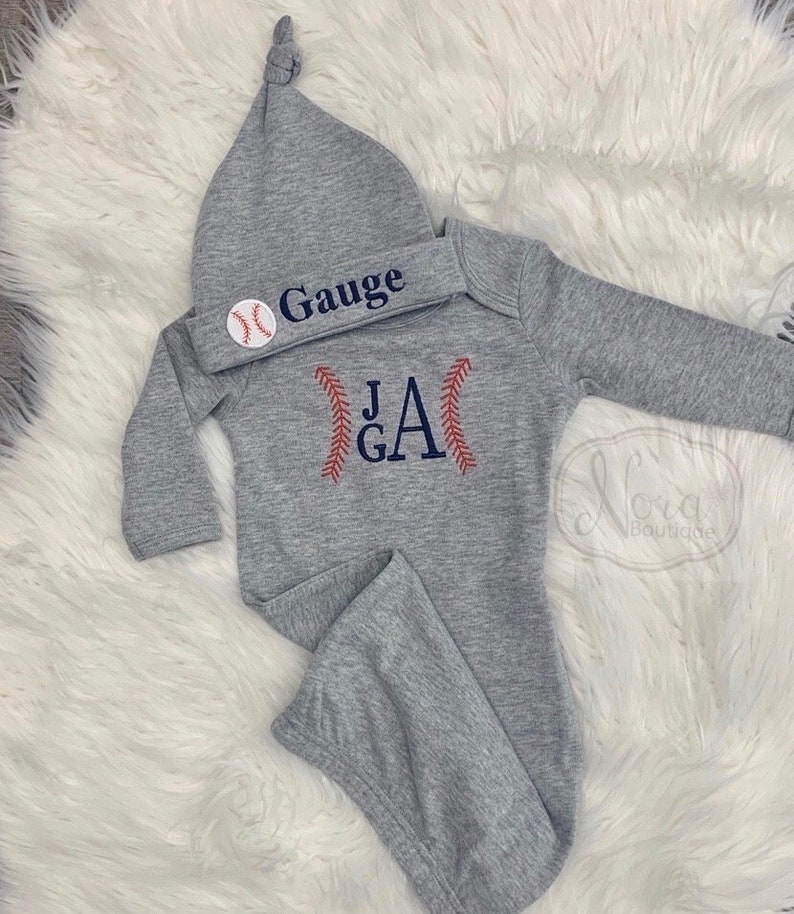 May include: Gray baby gown with long sleeves and a baseball-themed monogram with the initials "J.G.A." and red stitching. The gown is paired with a gray knit hat with a baseball graphic and the name "Gauge" embroidered on it.