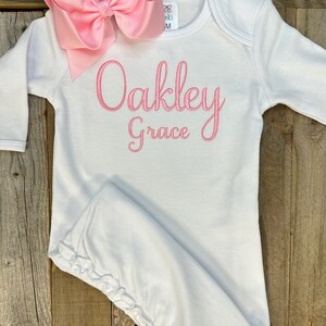 May include: A white baby gown with long sleeves and a pink bow. The gown has the name "Oakley Grace" embroidered in pink script. The bow is a large, light pink color and is placed at the top of the gown.