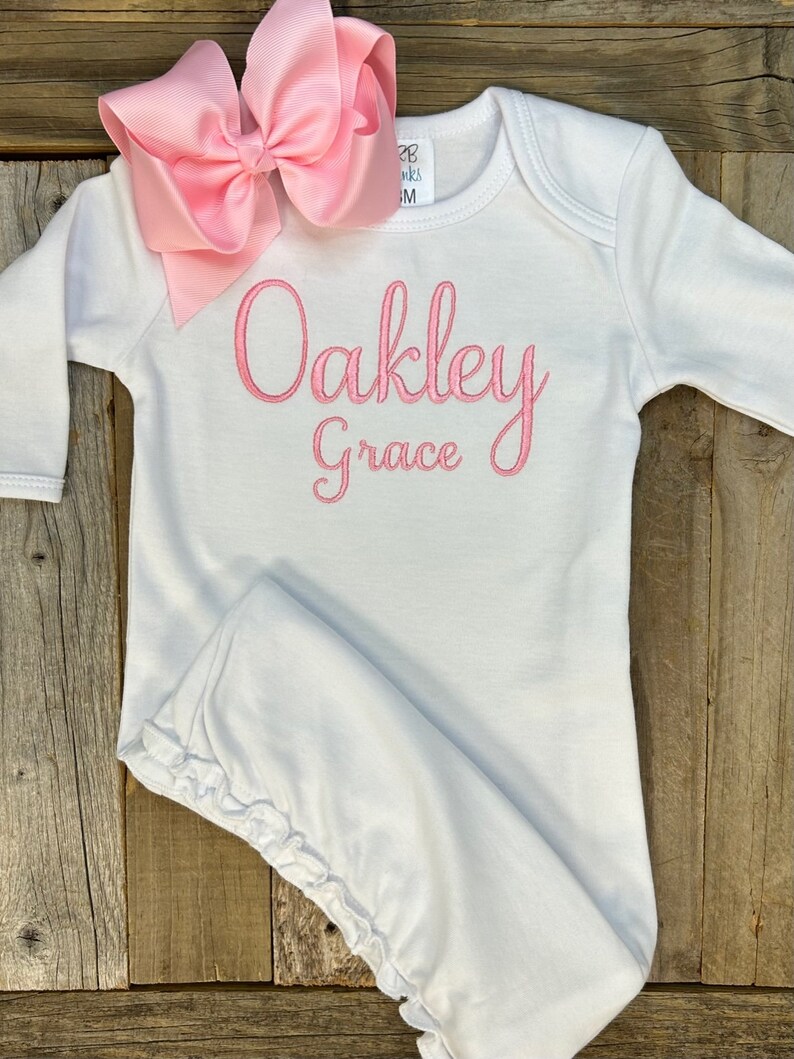 May include: A white baby gown with long sleeves and a pink bow. The gown has the name "Oakley Grace" embroidered in pink script. The bow is a large, light pink color and is placed at the top of the gown.