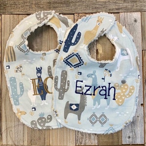 May include: Two blue baby bibs with a llama and cactus print. One bib is personalized with the name "Ezrah" and the other with the name "Eli".