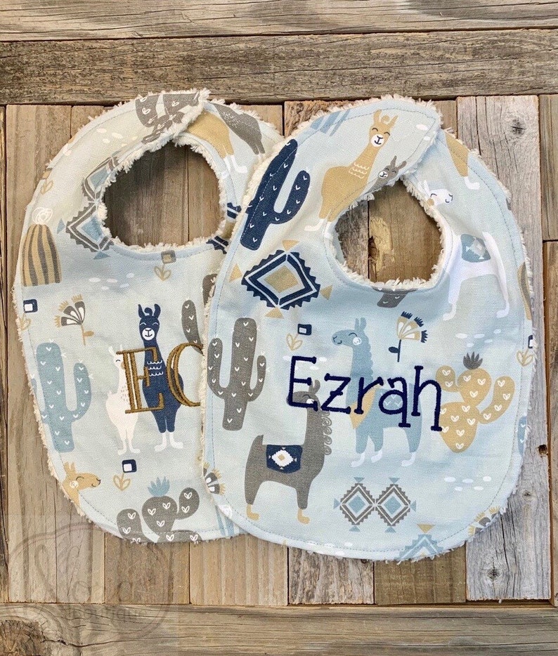 May include: Two blue baby bibs with a llama and cactus print. One bib is personalized with the name "Ezrah" and the other with the name "Eli".