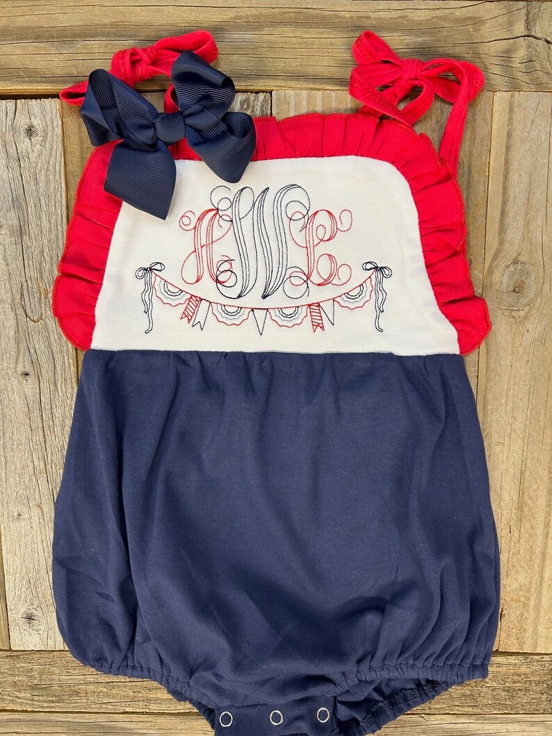 Puede incluir: Un pelele de beb&eacute; azul marino y rojo con un panel blanco que presenta un monograma y una guirnalda roja y blanca. Un lazo azul marino est&aacute; unido al pelele.