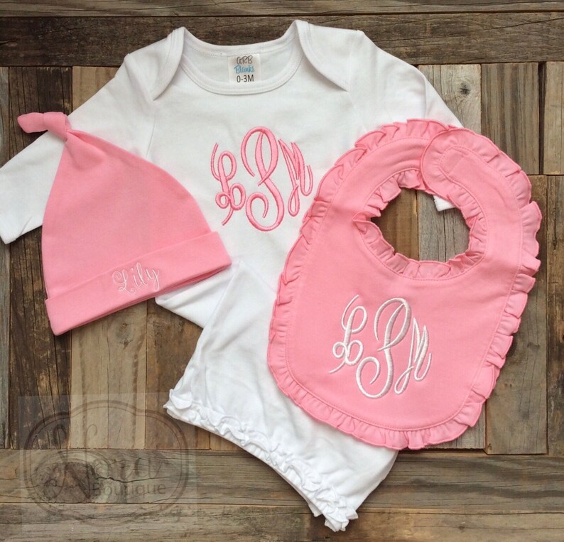 May include: A baby gift set featuring a white onesie, a pink bib with a ruffled edge, and a pink hat. The onesie and bib have embroidered initials, and the hat is embroidered with the name "Lily". The set is designed for a baby.