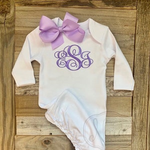 May include: A white baby gown with long sleeves and a lavender bow. The gown features a lavender monogram design on the chest. The gown is made of soft material and is designed for newborns.