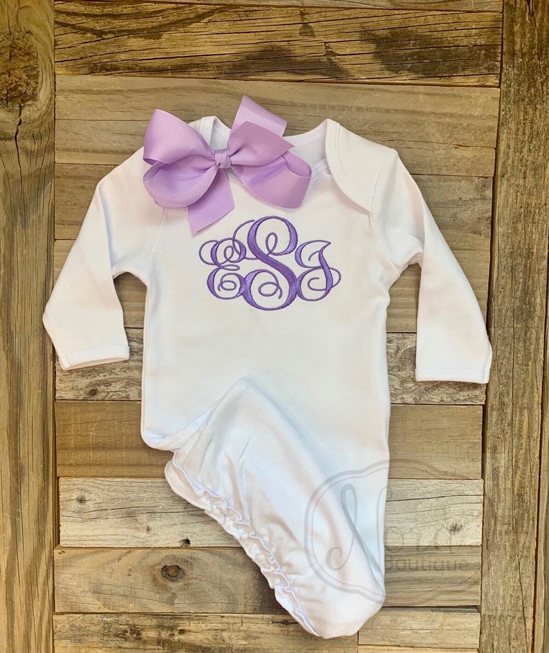 May include: A white baby gown with long sleeves and a lavender bow. The gown features a lavender monogram design on the chest. The gown is made of soft material and is designed for newborns.