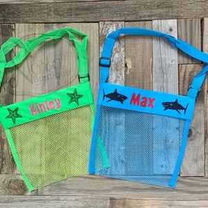 May include: Two mesh beach bags with adjustable straps. One bag is green with a pink name "Kinley" and a starfish. The other bag is blue with a red name "Max" and two black sharks.