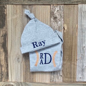 May include: Gray baby hat and onesie set with the name "Ray" embroidered on the hat and the initials "RAD" embroidered on the onesie. The onesie has orange stitching around the initials.