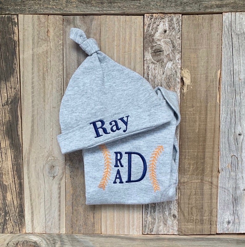 May include: Gray baby hat and onesie set with the name "Ray" embroidered on the hat and the initials "RAD" embroidered on the onesie. The onesie has orange stitching around the initials.