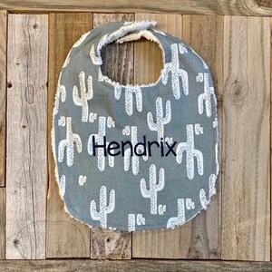 May include: A gray baby bib with a white cactus pattern and the name "Hendrix" embroidered in black.