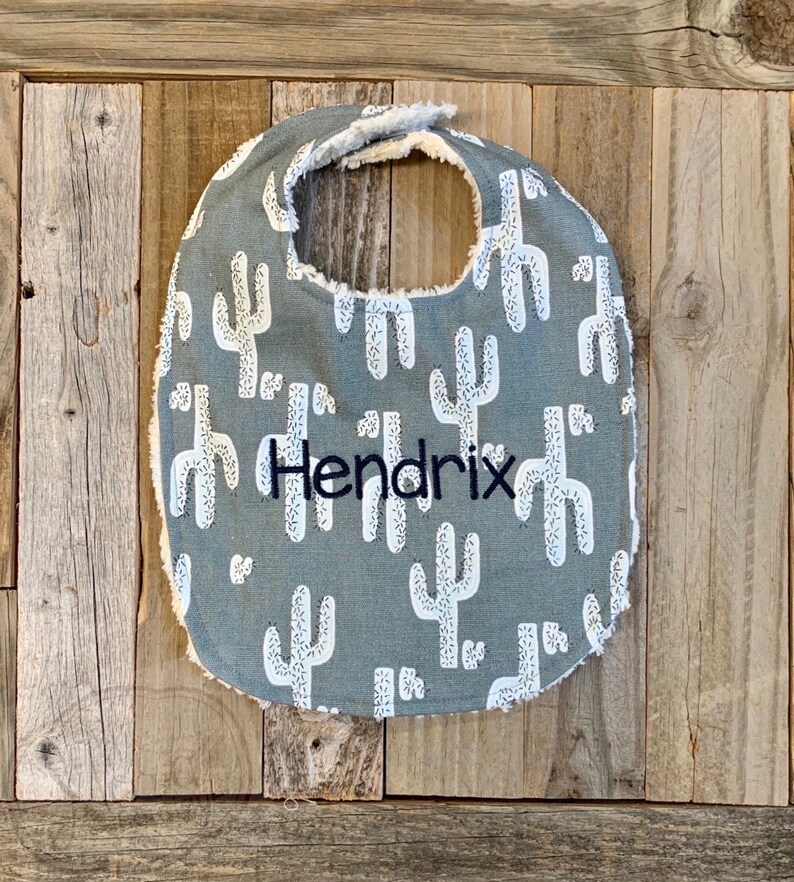 May include: A gray baby bib with a white cactus pattern and the name "Hendrix" embroidered in black.