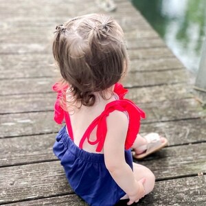 Puede incluir: Una ni&ntilde;a peque&ntilde;a con un mameluco azul y rojo con tirantes y volantes rojos. Est&aacute; sentada en un muelle de madera y mirando hacia el agua.