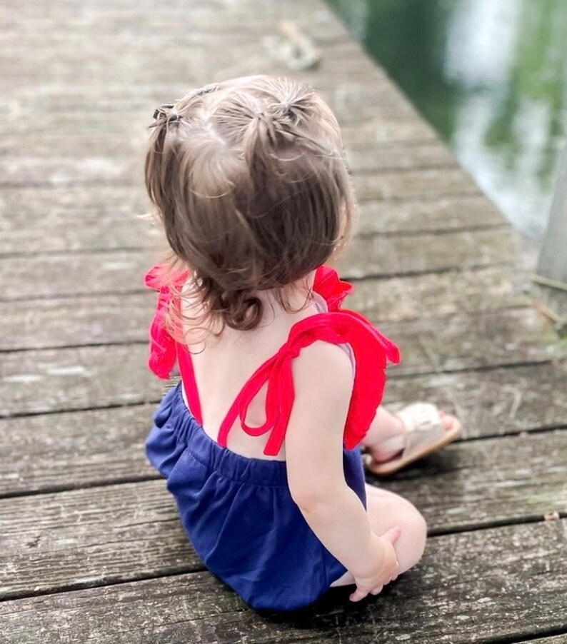 Puede incluir: Una ni&ntilde;a peque&ntilde;a con un mameluco azul y rojo con tirantes y volantes rojos. Est&aacute; sentada en un muelle de madera y mirando hacia el agua.