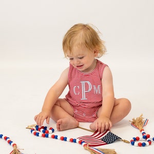 Op de afbeelding: Een peuter draagt een rood-wit gestreept, mouwloos rompertje met de letters "CPM" geborduurd op de voorkant en speelt met rood-wit-blauwe kralensnoeren en een kleine Amerikaanse vlag.