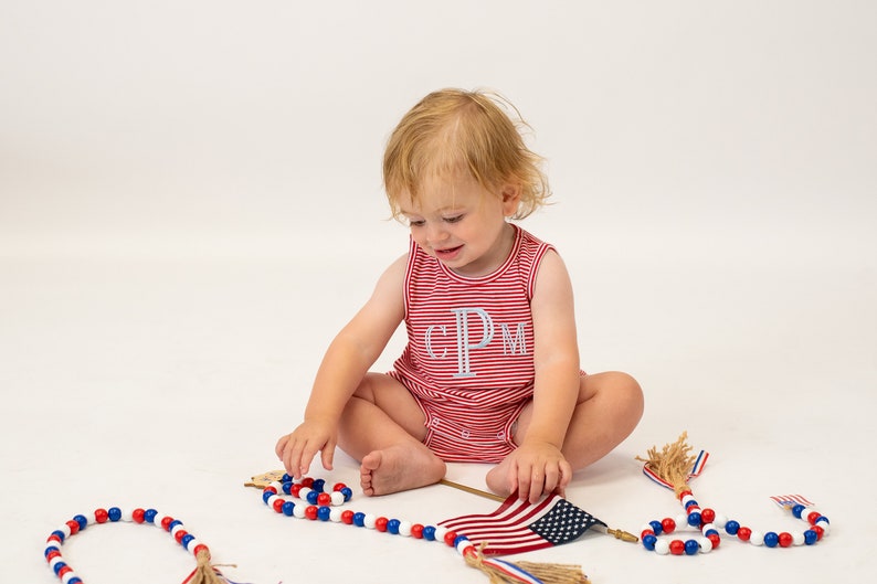 Op de afbeelding: Een peuter draagt een rood-wit gestreept, mouwloos rompertje met de letters "CPM" geborduurd op de voorkant en speelt met rood-wit-blauwe kralensnoeren en een kleine Amerikaanse vlag.