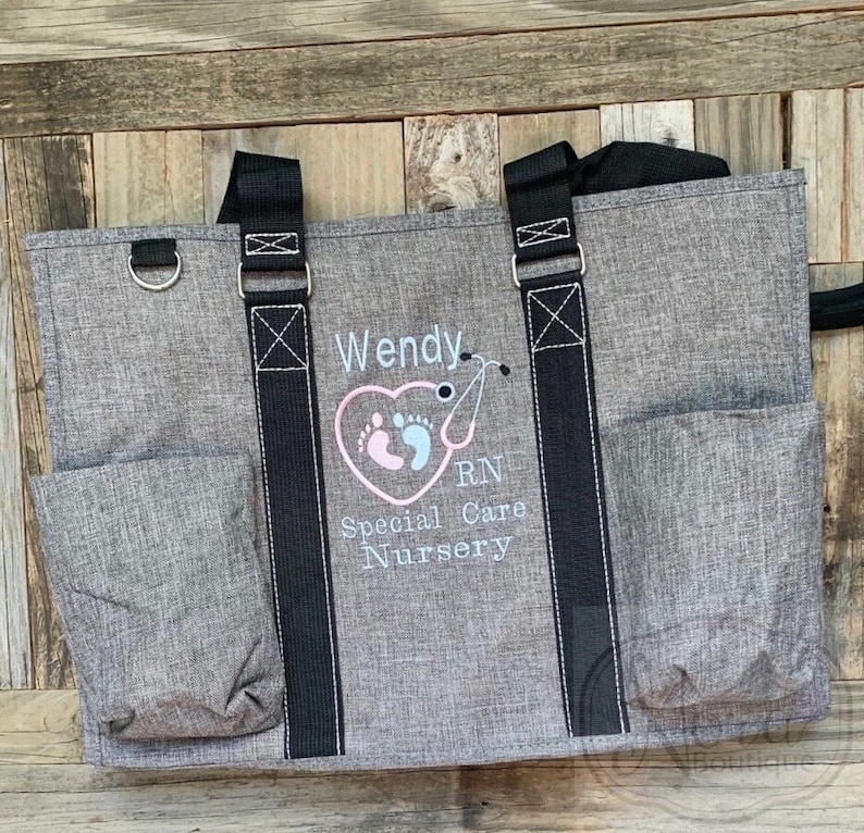 May include: A gray tote bag with black straps and a large embroidered design. The design features a pink heart with two footprints inside, a stethoscope, and the text "Wendy RN Special Care Nursery".