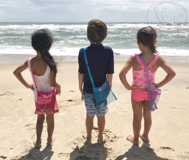 May include: Three children on a beach, wearing colorful swimsuits and carrying mesh bags with their names on them. The bags are pink, blue, and purple.