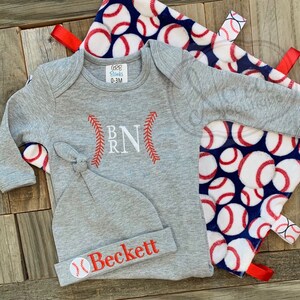 May include: A gray baby outfit with a baseball theme. The outfit includes a long-sleeved onesie with the letters "B R N" embroidered on the front, a gray knit hat with the name "Beckett" embroidered on the front, and a blue and white blanket with baseballs printed on it.