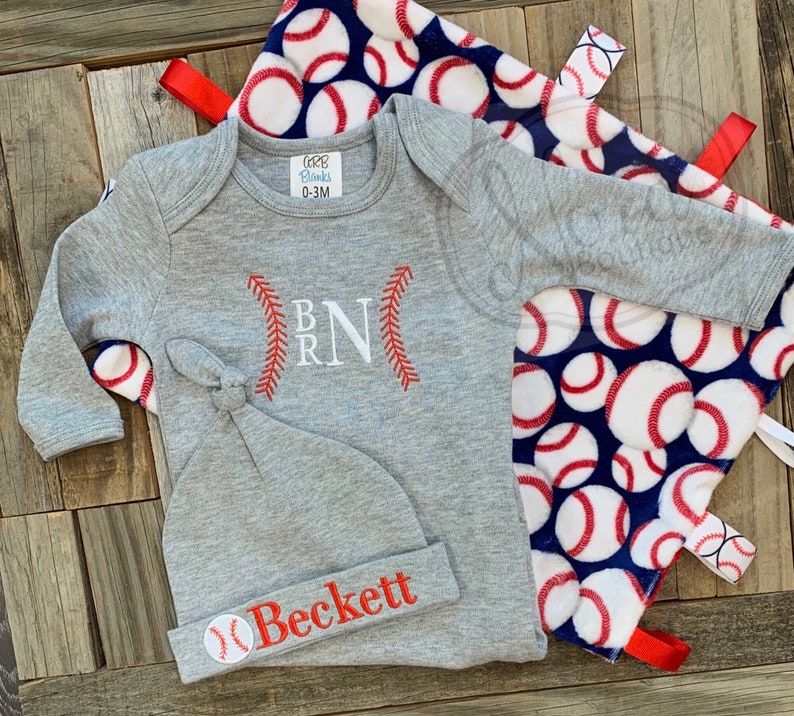 May include: A gray baby outfit with a baseball theme. The outfit includes a long-sleeved onesie with the letters "B R N" embroidered on the front, a gray knit hat with the name "Beckett" embroidered on the front, and a blue and white blanket with baseballs printed on it.
