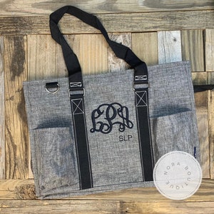 May include: A gray tote bag with black straps and a monogrammed design. The monogram is "SLP" in black embroidery. The bag has a zippered closure and a small pocket on the front.