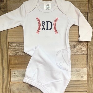 May include: White baby gown with long sleeves and a baseball-themed monogram. The monogram is "BAD" with the letters stitched in blue and the baseball stitching in red.