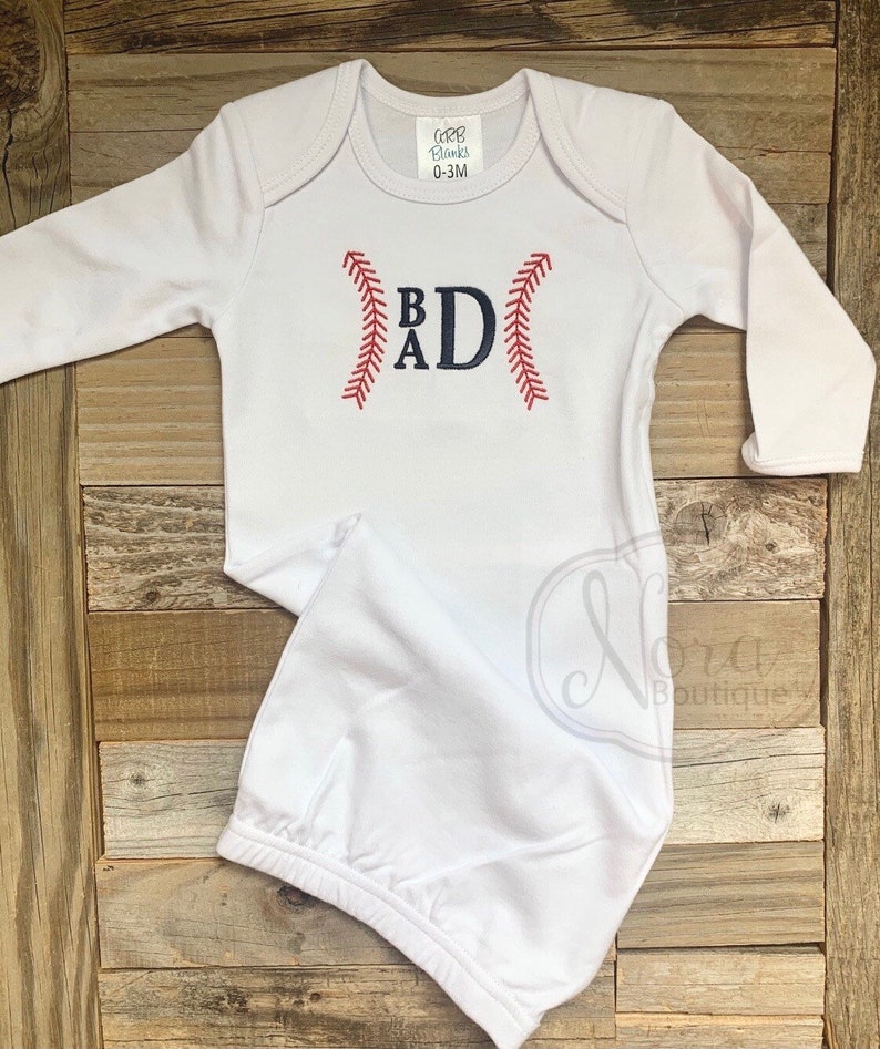 May include: White baby gown with long sleeves and a baseball-themed monogram. The monogram is "BAD" with the letters stitched in blue and the baseball stitching in red.