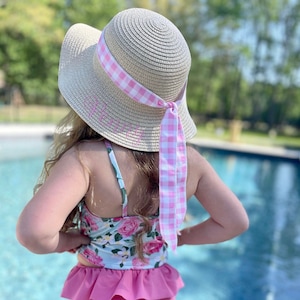 May include: A young girl wearing a pink and white gingham ribbon tied around a straw sun hat. The hat has a pink embroidered monogram. She is wearing a floral print swimsuit with a pink ruffle skirt.