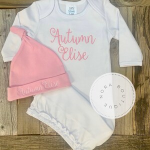 May include: A white baby outfit set with a matching pink hat. The set includes a long-sleeved onesie and a knotted hat, both embroidered with the name "Autumn Elise" in pink script. The outfit is displayed on a wooden surface.