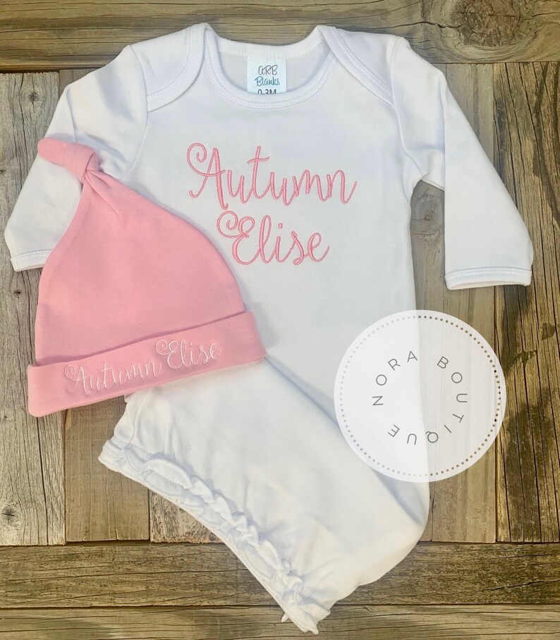 May include: A white baby outfit set with a matching pink hat. The set includes a long-sleeved onesie and a knotted hat, both embroidered with the name "Autumn Elise" in pink script. The outfit is displayed on a wooden surface.