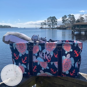 May include: A blue and white tote bag with a sea turtle and anchor pattern. The bag has a large pocket on the front with the monogram "MRJ" in white. The bag is sitting on a wooden dock overlooking a body of water.