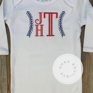 May include: White long-sleeve baby onesie with red and blue embroidered baseball-themed initials "JT" and a navy blue "Nora Boutique" label.