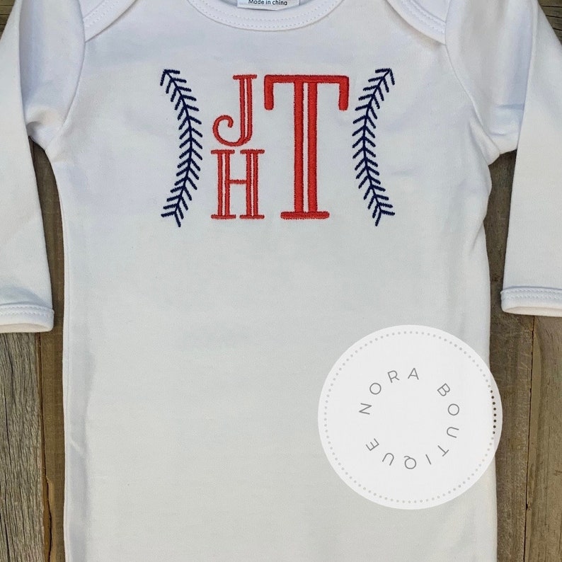 May include: White long-sleeve baby onesie with red and blue embroidered baseball-themed initials "JT" and a navy blue "Nora Boutique" label.