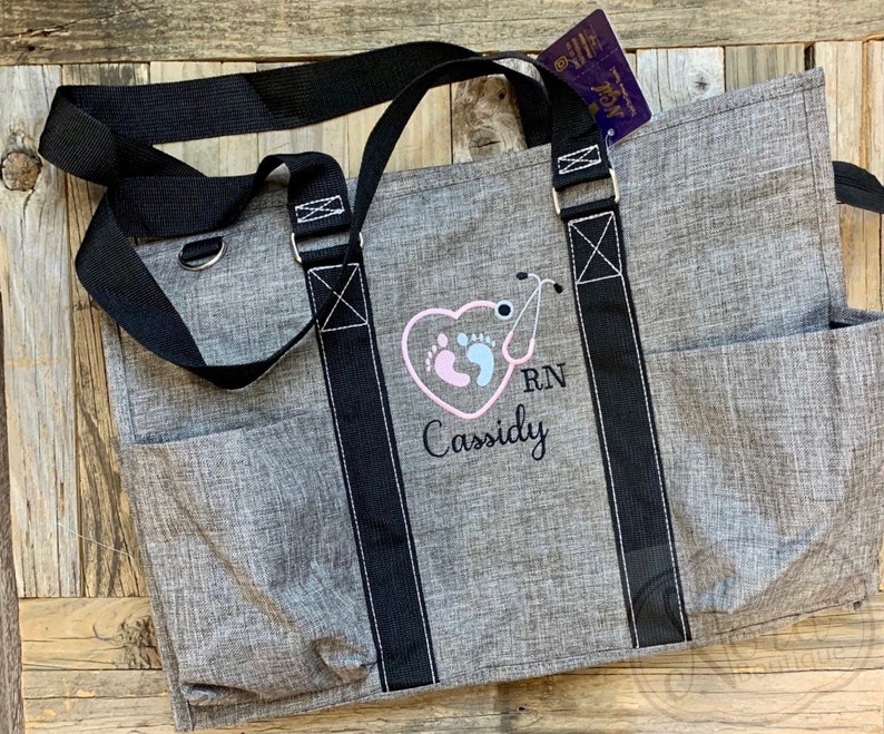 May include: Gray tote bag with black straps and a pocket on the side. The bag is embroidered with a pink heart, two footprints, a stethoscope, and the text "RN Cassidy".