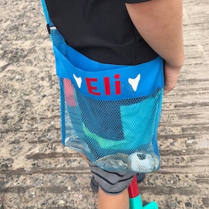 May include: A blue mesh beach bag with a red name tag that says "Eli" and white hearts on either side. The bag is worn over the shoulder and has a green shovel and other beach toys inside.