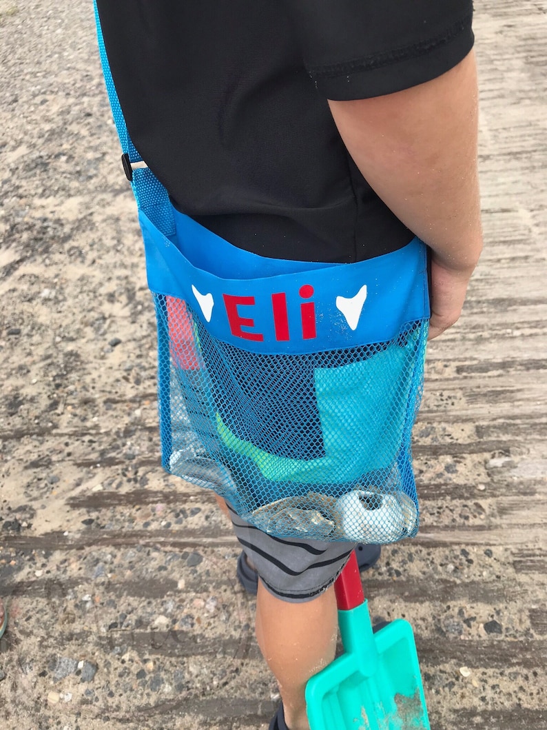 May include: A blue mesh beach bag with a red name tag that says "Eli" and white hearts on either side. The bag is worn over the shoulder and has a green shovel and other beach toys inside.