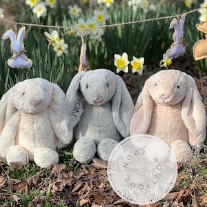 May include: Three plush bunny rabbits in cream, gray, and tan sit on the ground. The gray bunny has the name "Nora" embroidered on its ear. A garland of Easter bunny decorations hangs in the background, with daffodils.