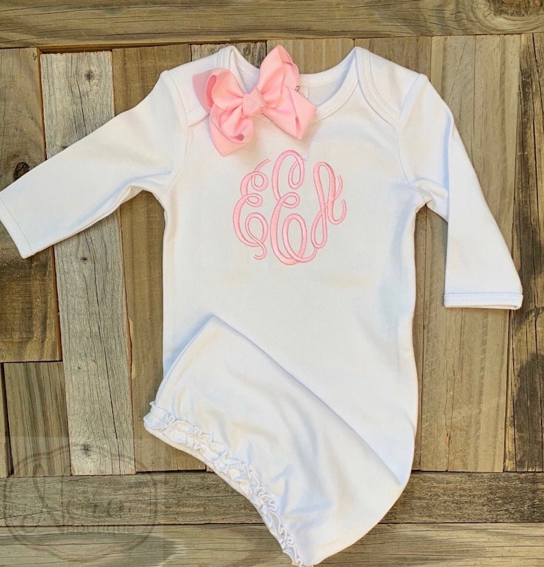 May include: White baby gown with long sleeves, a pink bow, and a pink monogram. The gown has a ruffled hem and is displayed on a wooden surface. The monogram features the letter "E".