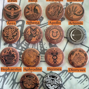 May include: Twelve round, orange-brown, ceramic buttons with black Greek god and goddess imagery. Each button features a different god or goddess, including Zeus, Hera, Poseidon, Demeter, Ares, Athena, Apollo, Artemis, Hephaestus, Aphrodite, Hermes, Dionysus, and two versions of Hades.