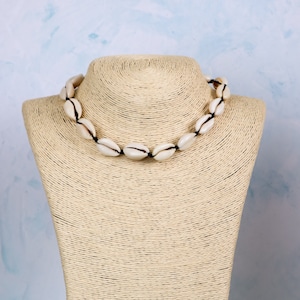 以下が含まれることがあります： ベージュのマネキンバストに白い貝殻のネックレス。ネックレスは、小さな白い貝殻を黒い糸でつないだものです。