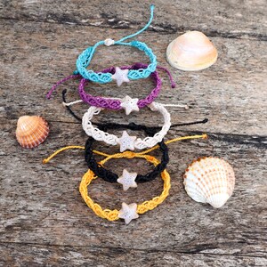 Puede incluir: Cinco pulseras de crochet de colores con abalorios de estrella de mar, dispuestas en una pila sobre una superficie de madera con conchas.