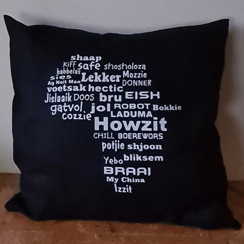 May include: Black throw pillow with white text that reads "shaap, kiff safe, shosholoza, babbelas, sies Lekker, Mozzie, Ag Noit Man, voetsok hectic, DONNER, Jislaaik Doos, bru EISH, gatvol, jol ROBOT Bokkie, LADUMA, Cozzie Howzit, CHILL BOEREWORS, potjie shjoon, Yebobliksem, BRAAI, My China, Izzit".
